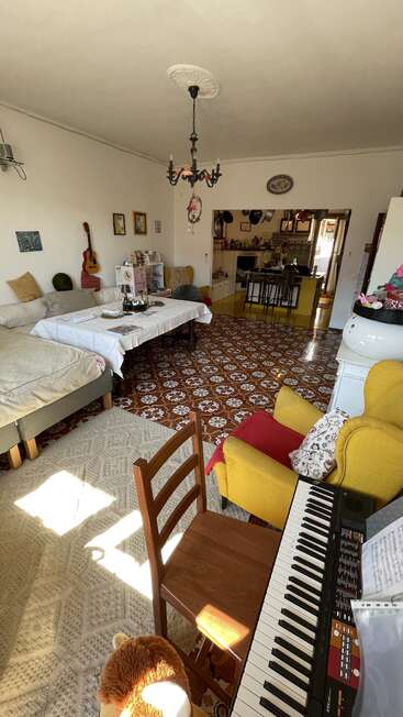 A cozy, eclectic living room with patterned floor, beds, a yellow armchair, keyboard, guitar, dining table, and visible kitchen. Sunlight brightens the space warmly.
