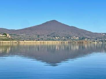 A mountain rises under a clear blue sky, perfectly reflected in the calm lake below. Sparse houses nestle along the shoreline, surrounded by natural beauty.
