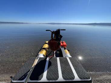 A small inflatable boat rests on a tranquil shoreline under a clear blue sky, with calm water stretching to distant hills, creating a peaceful, inviting scene.