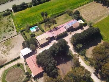 Cette image aérienne montre un grand domaine rural avec plusieurs bâtiments, une piscine, des panneaux solaires, des champs verdoyants, des arbres et des espaces paysagers, entourés de terres agricoles.