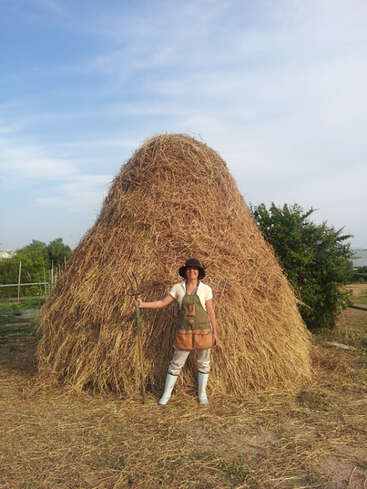 Une personne vêtue de vêtements de ferme et de bottes se tient devant une grande meule de foin, tenant une fourche, en plein air par une journée ensoleillée avec un ciel bleu.