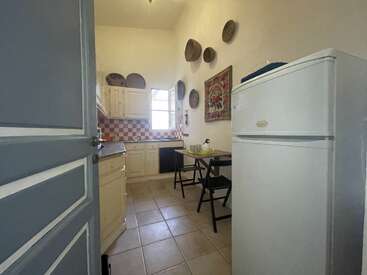 This is a cozy kitchen with cream cabinets, a checkered backsplash, a small dining table, wall decorations, a refrigerator, tiled floor, and open window.