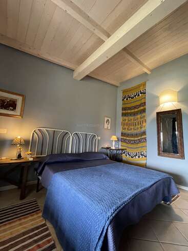 This cozy bedroom features a double bed with a blue blanket, wooden ceiling, striped rug, artistic tapestry, wall art, bedside lamps, and a rustic wooden desk.