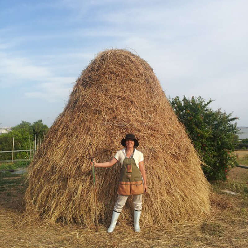 A person dressed in farm clothes and boots stands in front of a large haystack, holding a pitchfork, outdoors on a sunny day with blue sky.