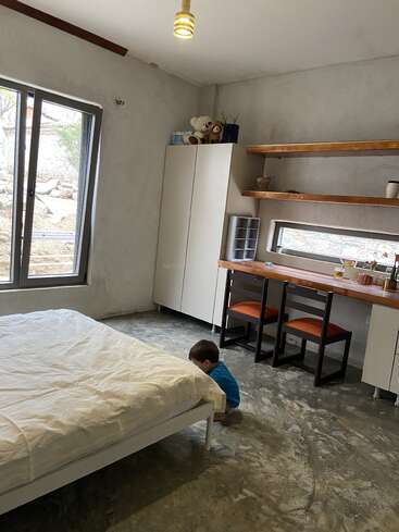 Um quarto arrumado com uma cama branca, janela grande, guarda-roupa, escrivaninha, cadeiras laranjas, prateleiras e uma criança pequena agachada perto da cama, olhando para baixo.