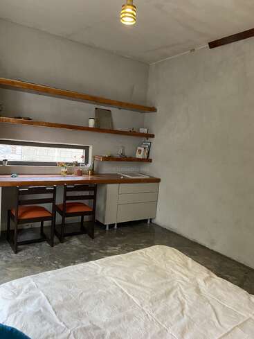 Esta imagem mostra um quarto minimalista com paredes e piso de concreto, uma cama simples, escrivaninha de madeira com duas cadeiras, prateleiras, iluminação suave e ambiente aconchegante.