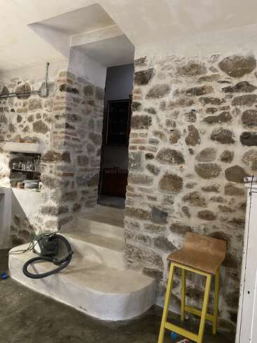 Esta imagem mostra um cômodo rústico com paredes de pedra, uma pequena escada, um banco de madeira com pernas amarelas, prateleiras com pratos e um aspirador de pó no chão.