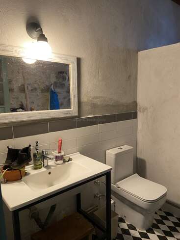 Esse banheiro tem uma pia branca com vários itens em cima, um espelho retangular com luz no teto, um vaso sanitário, paredes de azulejos e piso com padrão preto e branco.