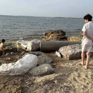 Uma mulher em pé em uma praia de areia, olhando para o oceano, cercada por grandes rochas, usando um vestido de cor clara e de costas para a câmera.