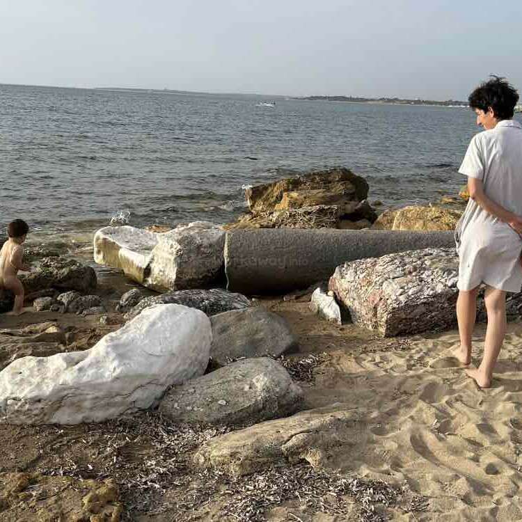 Uma mulher em pé em uma praia de areia, olhando para o oceano, cercada por grandes rochas, usando um vestido de cor clara e de costas para a câmera.