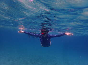 A person wearing a snorkel and a dark rash guard floats underwater, arms outstretched, surrounded by clear blue water and gentle ripples at the surface.