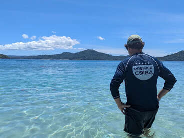 A man stands in clear, shallow water facing a scenic view of mountains and blue sky, wearing a "Premier Coiba" shirt and a green cap.