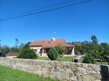 Uma charmosa casa de pedra com telhado de telhas vermelhas fica atrás de um muro de pedra rústico, cercada por vegetação, árvores e um céu azul claro e brilhante.