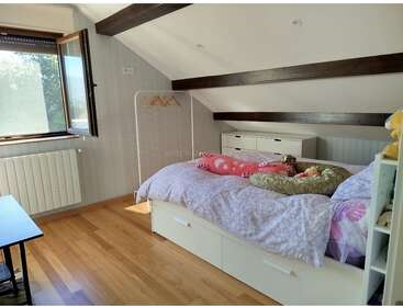 Esse quarto aconchegante no sótão apresenta um teto inclinado com vigas expostas, uma janela grande, piso de madeira, uma cama de solteiro coberta com brinquedos de pelúcia e móveis minimalistas.