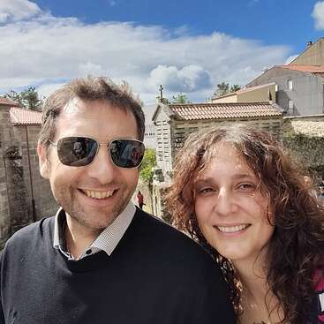 Um homem e uma mulher sorridentes posam ao ar livre em um dia ensolarado, com prédios antigos de pedra, telhados de telha e um céu claro e parcialmente nublado ao fundo.