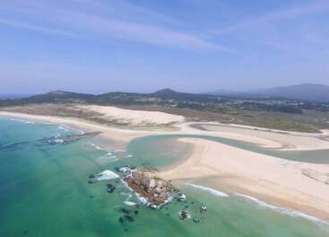 Uma vista aérea impressionante de uma praia intocada com água azul-turquesa, trechos de areia, um afloramento rochoso e colinas distantes sob um céu azul claro.