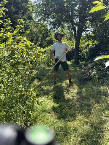 Un hombre está de pie en un jardín verde y soleado, sosteniendo una guadaña. Lleva un sombrero de ala ancha, una camisa blanca y pantalones cortos, rodeado de una densa vegetación.