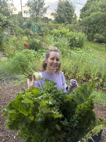 Una mujer sonriente posa en un frondoso jardín, sosteniendo hinojo, colinabo y un gran manojo de col rizada recién cosechados, rodeada de una vegetación exuberante.
