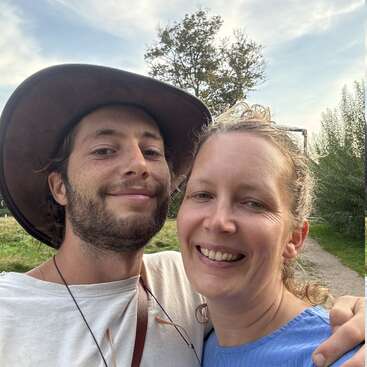 Um casal sorridente está bem juntinho ao ar livre; o homem usa um chapéu de abas largas e camisa branca, e a mulher está vestida de azul, aproveitando um dia ensolarado e repleto de natureza.