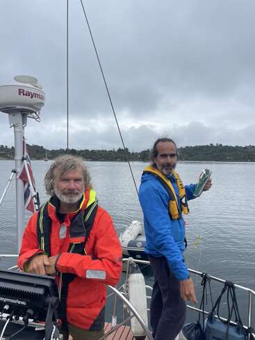 Zwei Männer stehen auf einem Segelboot, gekleidet in bunte Jacken und Schwimmwesten. Das Wetter ist bewölkt, und sie sind von ruhigem Wasser und Bäumen umgeben.