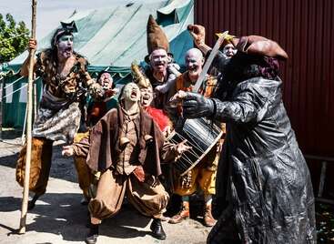 Eine lebhafte Gruppe von Menschen in aufwendigen Fantasiekostümen, mit Gesichtsbemalung und Masken posiert begeistert im Freien, einige schreien oder gestikulieren, im Hintergrund ist ein Zelt zu sehen.