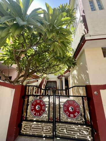 Ein Wohnhaus mit cremefarbenen Wänden, roten Bordüren und einem dekorativen Tor mit Yin-Yang-Symbolen. Ein großer, belaubter Baum spendet Schatten und verstärkt die einladende Atmosphäre.