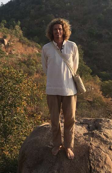 Ein barfüßiger Mann mit lockigem Haar steht auf einem Felsen, trägt ein helles Hemd und eine Hose und hat eine Tasche dabei. Hügel und Bäume bilden den Hintergrund.