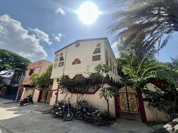 Ein strahlender Sonnentag mit einem zweistöckigen, cremefarbenen Haus mit roter Umrandung, üppigem Grün, draußen geparkten Motorrädern und einem klaren blauen Himmel mit vereinzelten Wolken.