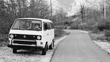 Una furgoneta blanca vintage está aparcada junto a una carretera sinuosa en un paisaje rural invernal. Árboles desnudos rodean la zona y un perro está sentado dentro.