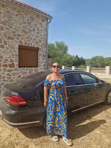 Una mujer con gafas de sol y un vestido colorido azul y amarillo posa con confianza junto a un coche negro, delante de una casa de piedra bajo un cielo soleado.