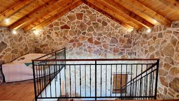Este acogedor loft cuenta con paredes rústicas de piedra, techos de madera con vigas a la vista, iluminación cálida, barandillas metálicas, suelos de madera y una bañera de hidromasaje en esquina para relajarse.