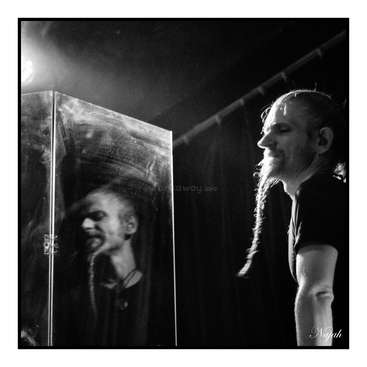 The image is a black-and-white photograph of two men, one with a long braid, standing in front of a glass door, with a spotlight shining down on them.