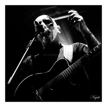 The image depicts a man playing an acoustic guitar and singing into a microphone, with a spotlight shining down on him in a dark setting.