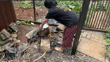 A person is cooking outdoors over an open fire using a large pot, surrounded by rocks and twigs, wearing slippers and casual clothing in a fenced backyard.
