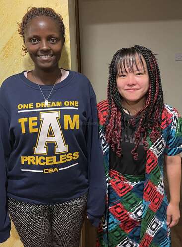 Two women stand indoors smiling at the camera; one wears a dark blue sweater with yellow writing, the other wears colorful patterned clothes with braided hair.