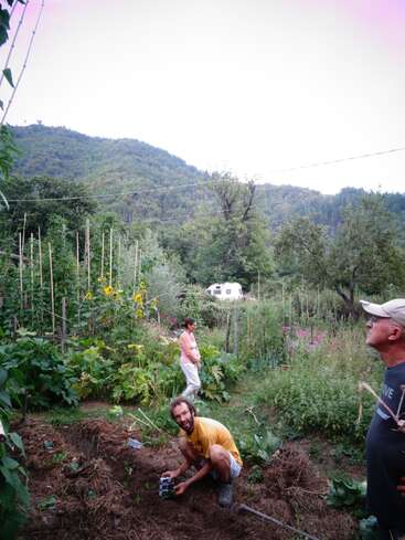 Three people are in a lush vegetable garden surrounded by greenery and mountains. One person kneels, smiling while gardening, while the others stand observing and walking.