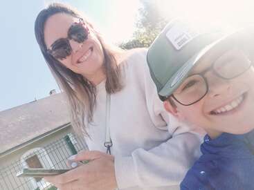 Uma mulher usando óculos escuros e um menino de óculos e boné estão sorrindo ao ar livre. O sol está brilhando intensamente e eles parecem felizes juntos.