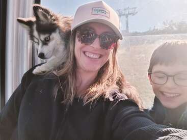 Uma mulher usando óculos escuros e chapéu sorri enquanto está sentada com um menino feliz e um cachorro preto e branco fofo pousado em seu ombro dentro de um teleférico.