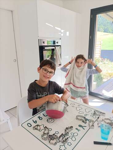 Duas crianças estão em uma cozinha bem iluminada. Um menino mistura ingredientes em uma tigela, enquanto uma menina brinca com uma toalha na cabeça, fazendo orelhas de coelho.