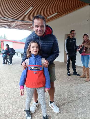 Um homem e uma jovem posam juntos do lado de fora, sob um telhado de madeira. A garota usa uma jaqueta colorida e há pessoas visíveis ao fundo.