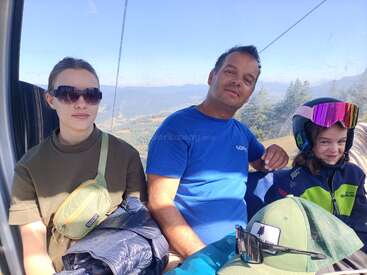 Três pessoas sentadas dentro de um teleférico, cercadas por montanhas panorâmicas. Uma delas usa óculos escuros, a outra usa uma camisa azul e a terceira usa capacete e jaqueta de esqui.