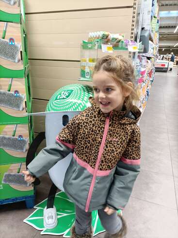 Uma jovem sorridente, vestindo uma jaqueta com estampa de leopardo, está em um corredor de uma loja perto de um mostruário de Tic Tac, cercada por prateleiras e caixas de produtos de limpeza.