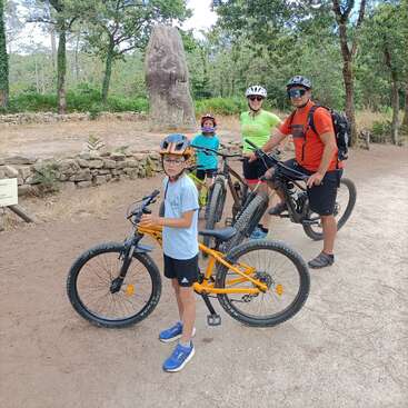 Quatro pessoas, usando capacetes e equipamentos de ciclismo, posam com suas bicicletas em um caminho de terra em uma área arborizada, perto de um grande monumento de pedra.