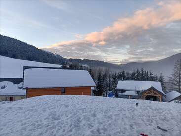Casas cobertas de neve aninham-se em uma serena paisagem montanhosa ao pôr do sol. Os pinheiros cercam as cabanas aconchegantes, enquanto o céu brilha com suaves nuvens rosa e laranja.