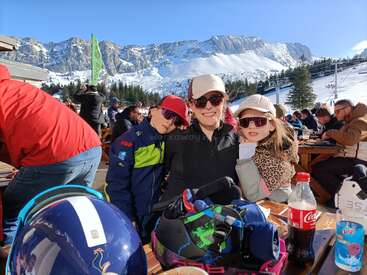 Uma mulher e duas crianças, todas usando óculos escuros e roupas de inverno, sorriem em um café de uma estação de esqui. Montanhas nevadas e esquiadores são visíveis ao fundo.