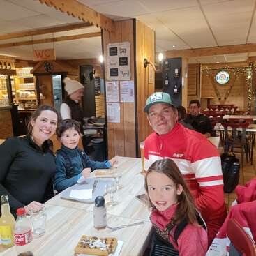 A smiling family of four sits at a wooden table in a cozy restaurant, enjoying waffles and drinks. Warm lighting and rustic decor create a welcoming atmosphere.