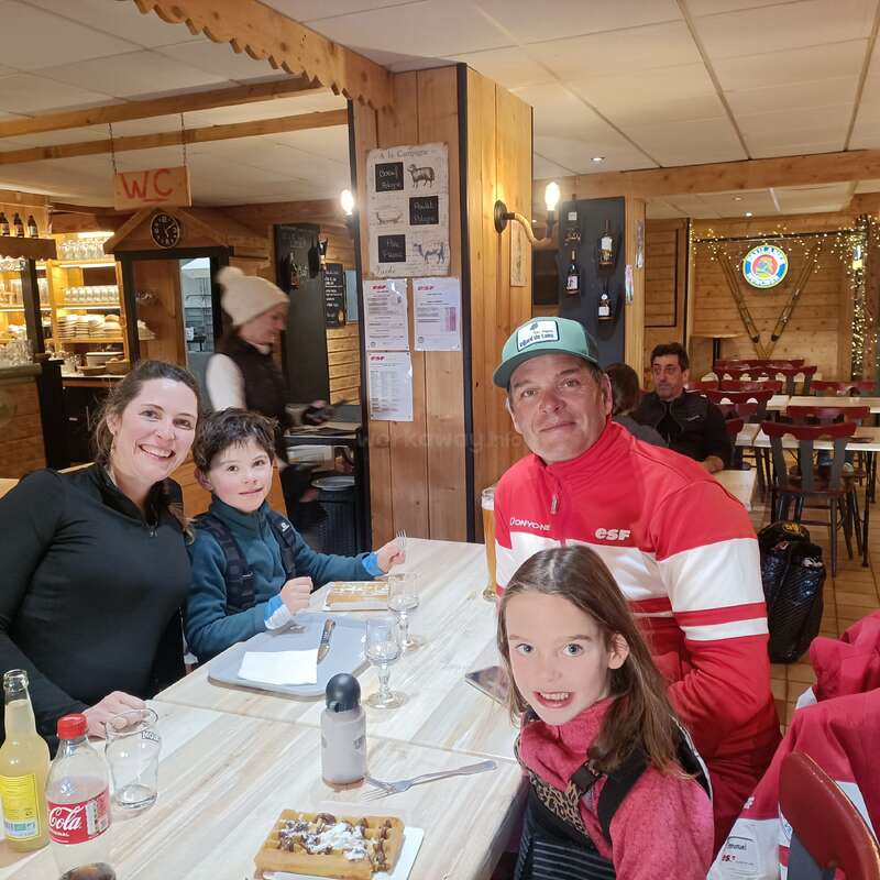 Uma família sorridente de quatro pessoas senta-se em uma mesa de madeira em um restaurante aconchegante, saboreando waffles e bebidas. A iluminação quente e a decoração rústica criam uma atmosfera acolhedora.