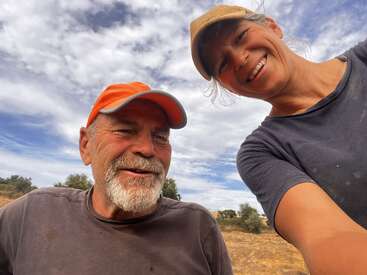 Un homme et une femme souriants, tous deux coiffés d'un chapeau, prennent un selfie en plein air sous un ciel partiellement nuageux. Ils ont l'air heureux, peut-être en train de vivre une aventure à la campagne.