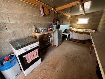 Cette image montre une petite pièce rustique avec une kitchenette, des appareils électroménagers et un lit bien rangé dans un coin. Les murs sont des blocs de béton non finis.