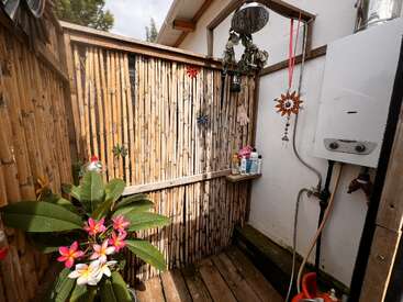 Voici une douche extérieure en bambou avec un sol en bois, des fleurs de plumeria, des objets décoratifs, un chauffe-eau, des bouteilles de shampoing et des ornements suspendus. La lumière naturelle l'éclaire.